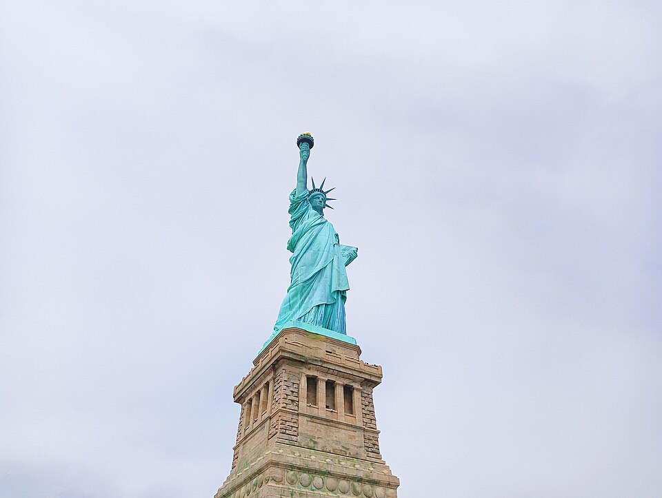 The Statue of Liberty viewed from below, holding a torch and tablet above its stone pedestal under a pale sky.